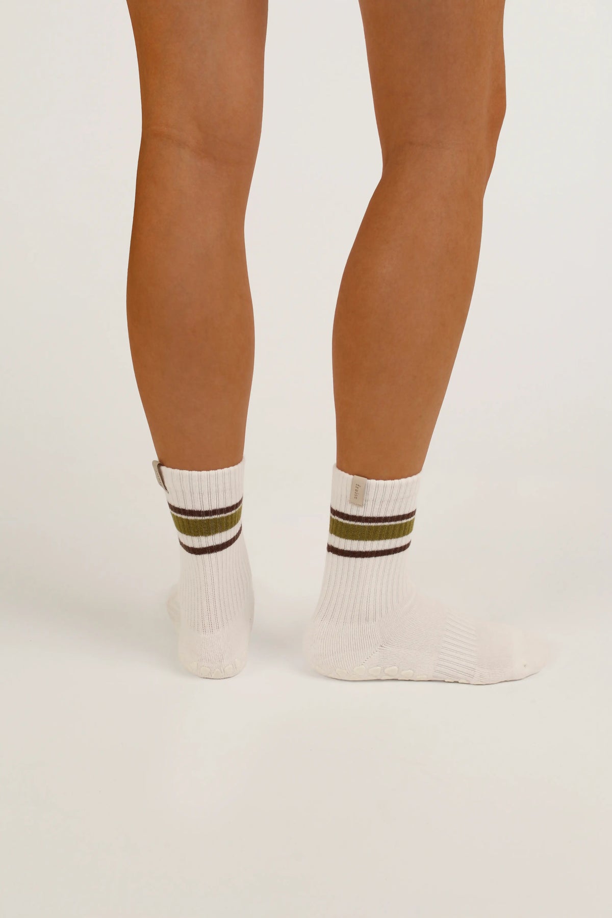 White pilates crew grip socks with olive green and dark brown stripes near the cuff, shown on a model's feet against a plain light background.