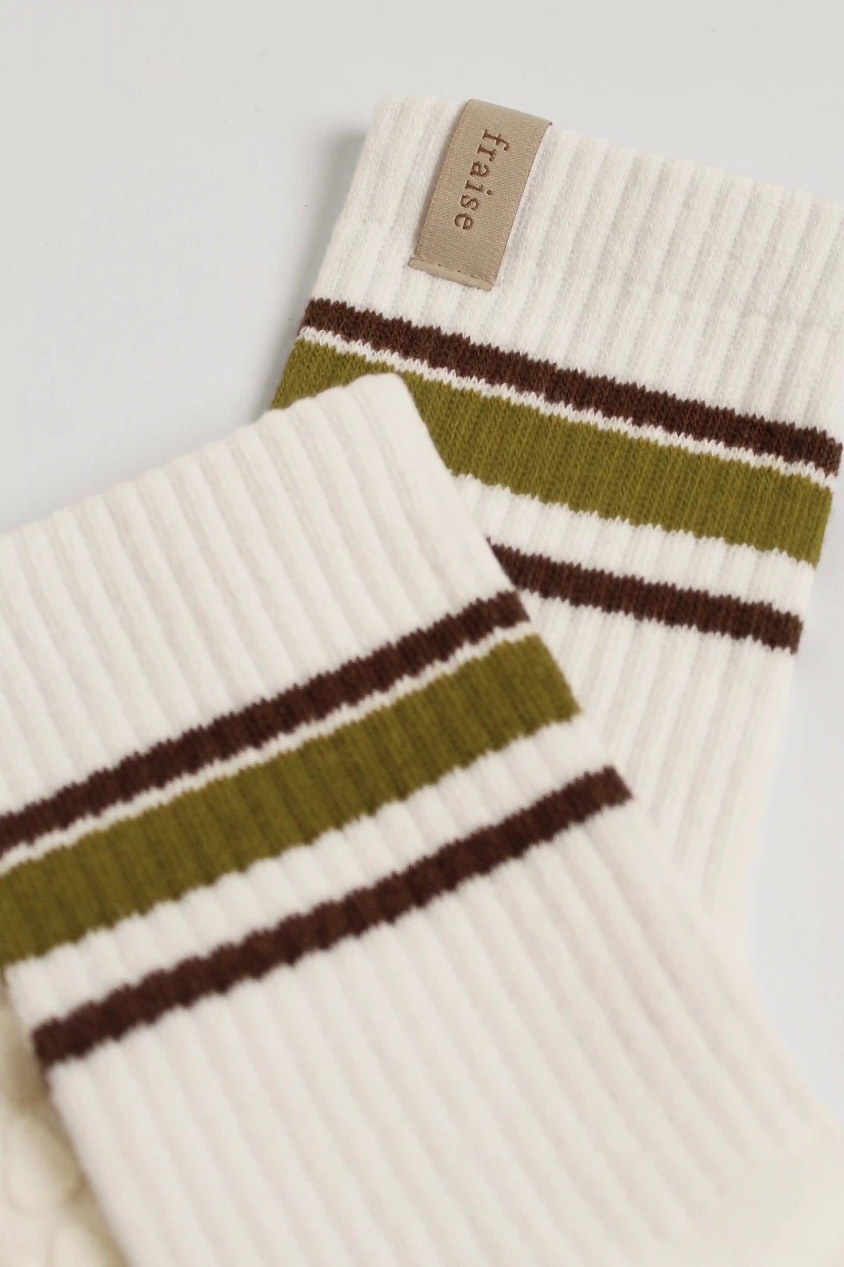 A close-up of a pair of ribbed white grip socks with olive green and dark brown stripes, featuring a beige 'fraise' brand tag.
