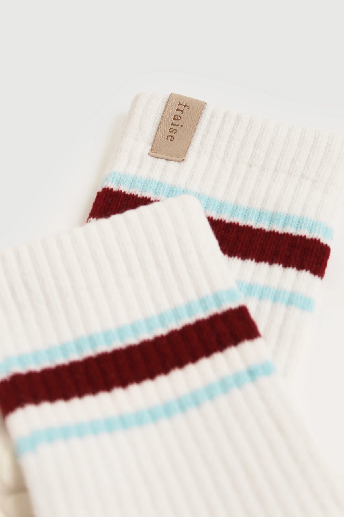 A close-up of a pair of white ribbed knit pilates socks with horizontal stripes in light blue and dark burgundy, featuring a small tan tab that reads "Fraise".