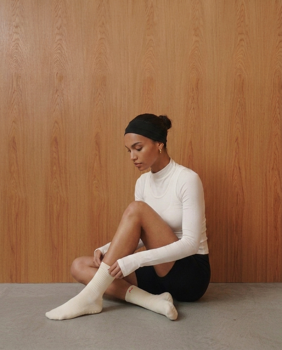 A woman sits on a concrete floor against a light wood-paneled wall, wearing a white long-sleeve mock-neck top, black shorts, and off-white ribbed pilates grip socks with "fraise" embroidered on the cuff.