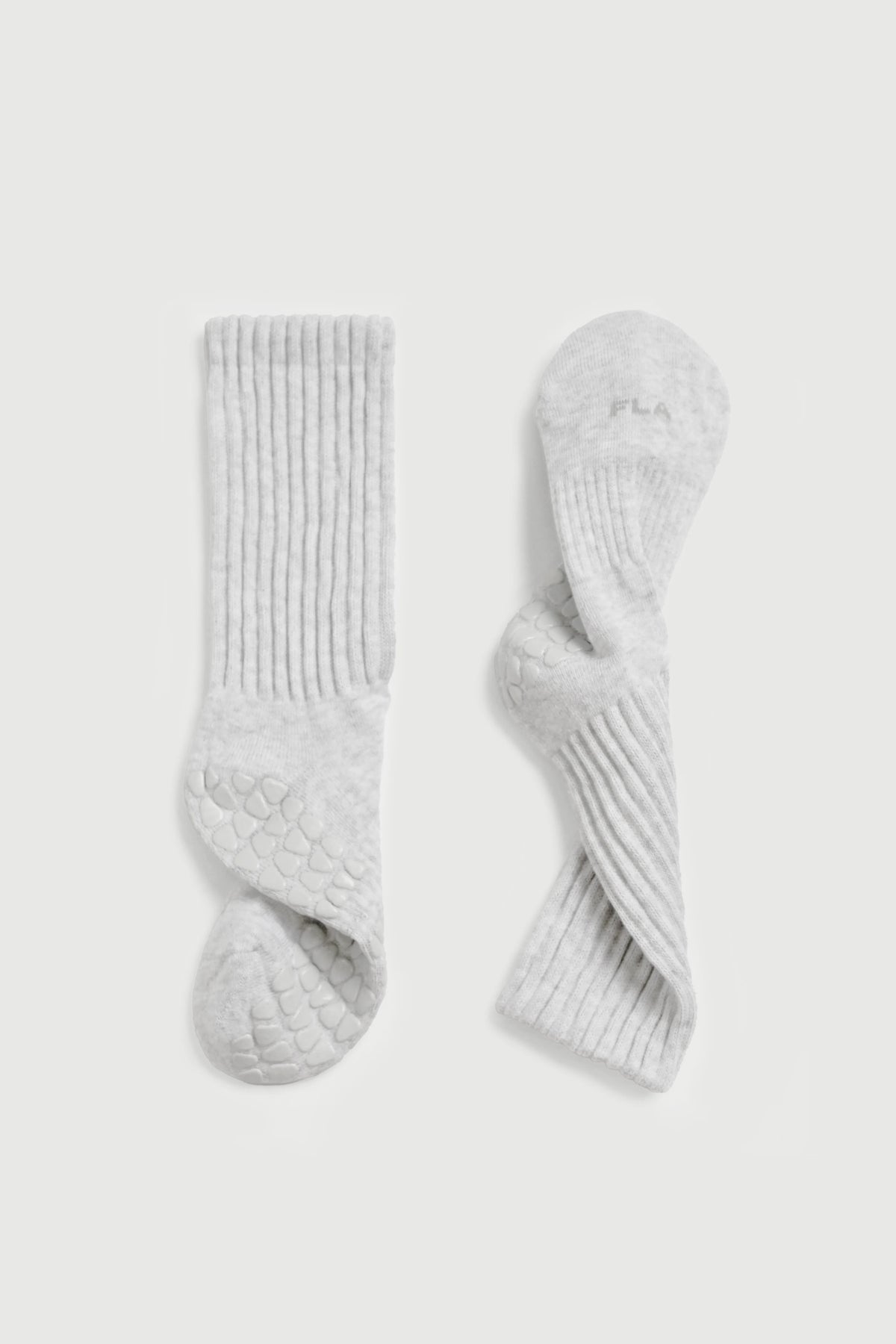A pair of light gray ribbed pilates socks with textured grip soles, shown flat on a light gray background. One sock is upright showing the ribbed leg and textured sole, and the other is curled to show the grip pattern on the foot.