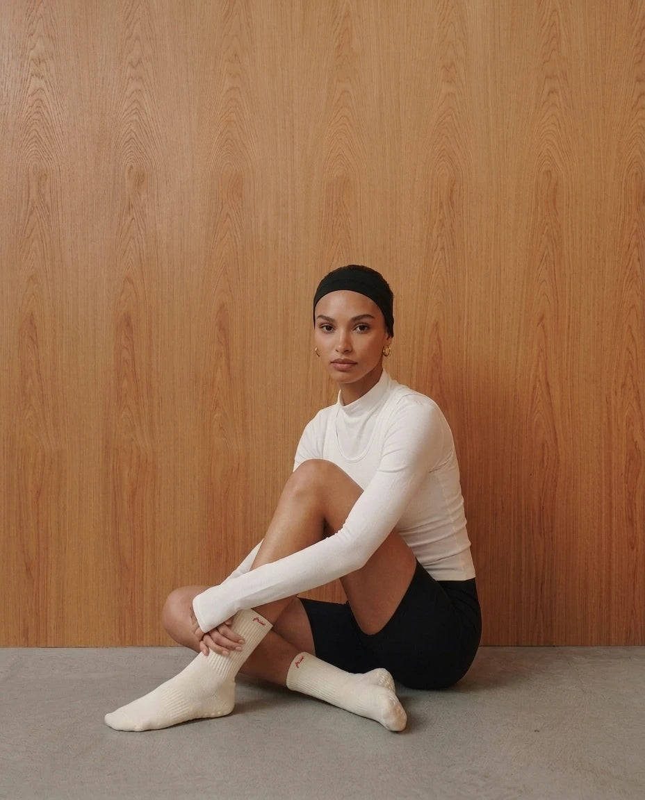 A woman sits cross-legged against a light wood-paneled wall, wearing a fitted white mock-neck long-sleeve top, black shorts, and off-white pilates crew grip socks with "Fraise" embroidered on them. 